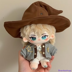 May include: A plush doll with blonde hair wearing a brown witch hat, a gray jacket with gold accents, and white pants. The doll has green eyes and a cute expression.