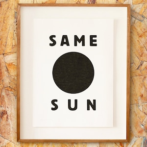 May include: A framed art print with the words "SAME SUN" in bold black letters. A large black circle is centered between the words. The print is in a white mat and a light brown wooden frame.