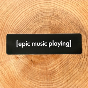 May include: A black rectangular sticker with the text "[epic music playing]" in white. The sticker is placed on a light brown, wooden surface with a circular grain pattern. The sticker's design is simple and modern.