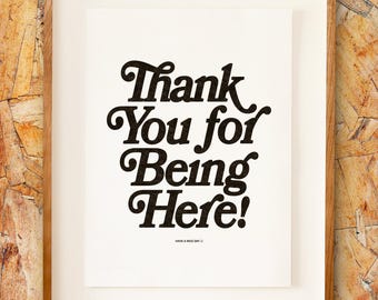 Puede incluir: Un cartel enmarcado con la frase "Thank You for Being Here!" en negro, con una fuente retro. La impresión está sobre un fondo blanco, dentro de un marco de madera marrón claro. La frase "Have a nice day" está en la parte inferior.