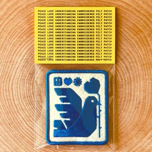 Puede incluir: Parche de fieltro bordado con un diseño de paloma azul que lleva un tallo con un corazón y otras formas. El parche está en una funda de plástico transparente. El texto "PEACE LOVE UNDERSTANDING EMBROIDERED FELT PATCH" está impreso en una etiqueta amarilla.