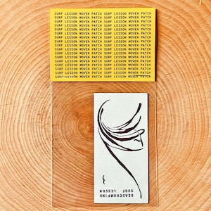 May include: A woven patch with a minimalist design of a person's face and the words "BEAUCHAMPING SURF LESSON". The patch is in a clear plastic sleeve, with a yellow header repeating "SURF LESSON WOVEN PATCH". The background is a wooden surface.