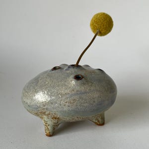 May include: A ceramic bud vase with a light blue and silver glaze, featuring three small holes for stems and three short legs. A single yellow pom-pom flower is inserted into one of the holes, creating a unique floral arrangement.
