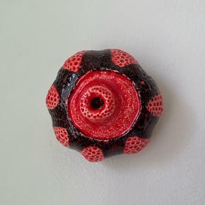May include: A ceramic sculpture with a central red, circular design and a dark, textured exterior. The outer surface features a pattern of red dots against a dark background, creating a striking contrast. The sculpture has a unique, organic shape.