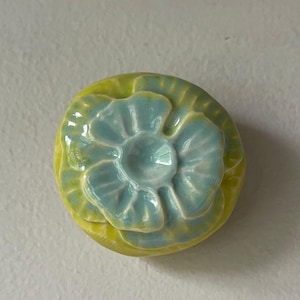 May include: A ceramic flower-shaped object with a light blue center and yellow-green outer petals. The handmade piece has a glossy finish and is likely a decorative item or a small dish.