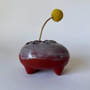 May include: A small, handmade ceramic vase in a rounded shape, with a deep red base and a gray-white top. The vase has three small legs and holds a single dried flower with a yellow, spherical head.