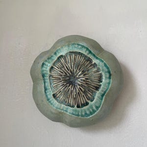 May include: A circular ceramic wall art piece with a scalloped edge. The outer rim is a matte sage green, transitioning into a vibrant teal band that surrounds a dark, textured center featuring radiating, carved lines resembling a flower.