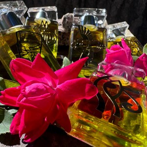 May include: A group of five clear glass bottles with gold and black labels, filled with a yellow liquid. The bottles are arranged in a cluster, with a pink rose in the foreground.
