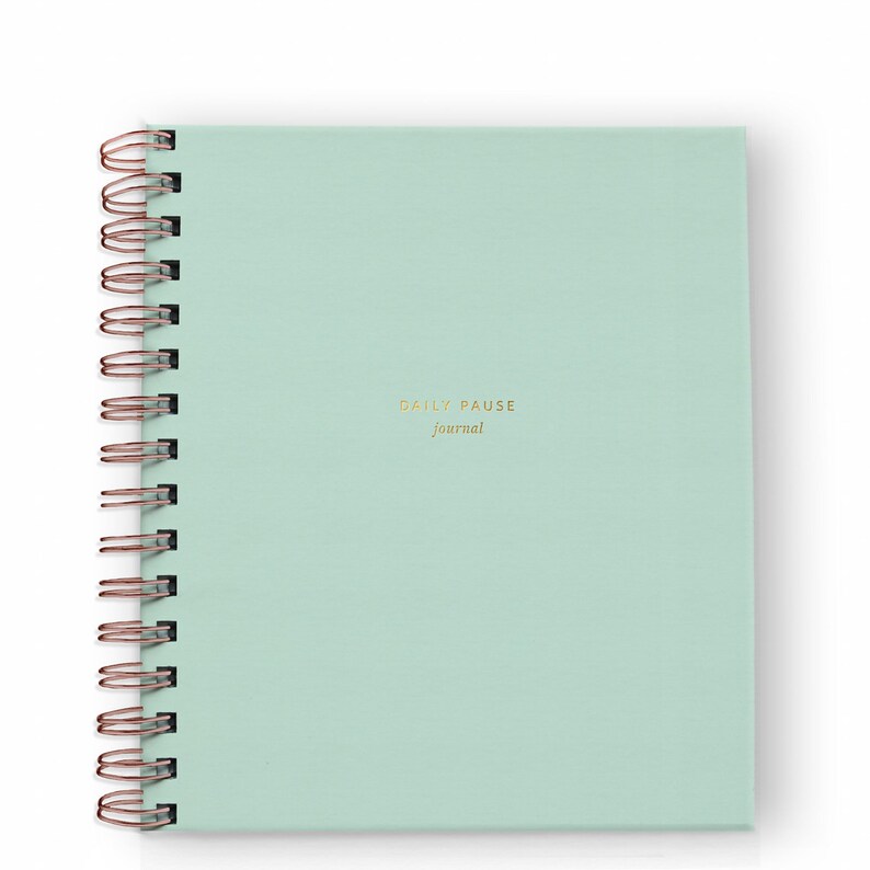 May include: A mint green spiral-bound journal with the words "DAILY PAUSE journal" in gold lettering. The spiral binding is rose gold. The journal is square and sits on a white background.