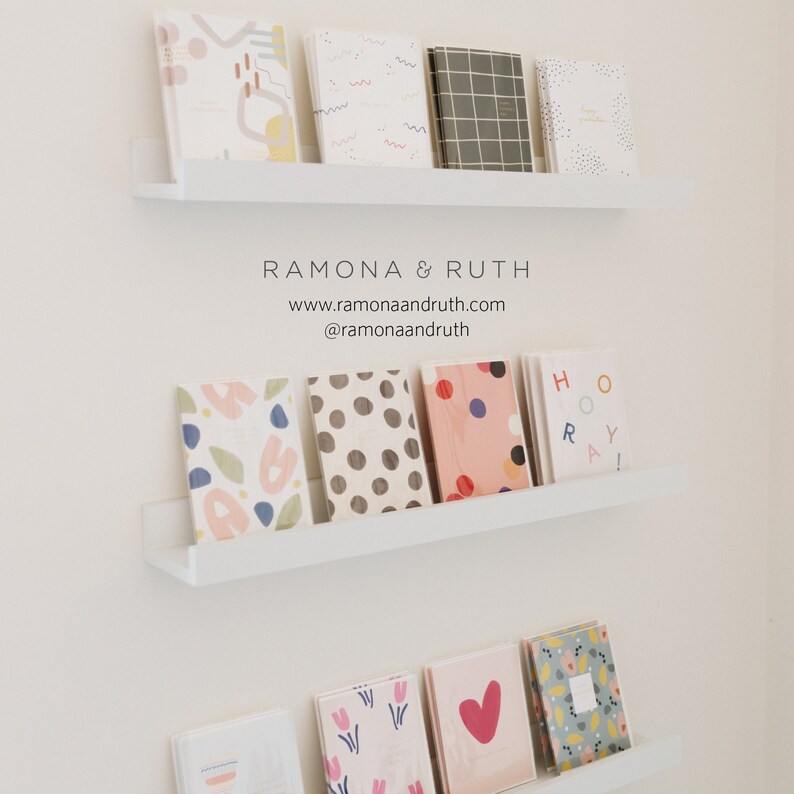 May include: A white shelf with a variety of colorful notebooks and journals. The notebooks have different patterns, including polka dots, stripes, and abstract designs. The shelf is labeled "Ramona & Ruth" with the website address and social media handle.