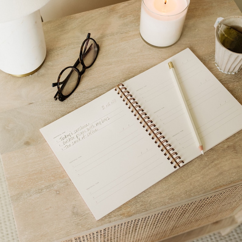 May include: A white spiral bound notebook with a pencil on top of it. The notebook has the date 5/12/20 written on the top page. The text "Today's sunshine - Outdoor picnic with my boys - The smell of coffee" is written on the page.