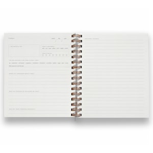May include: Open journal with lined pages and a spiral binding. The left page features prompts for gratitude, feelings, and daily experiences. The right page offers space for additional thoughts. The journal is designed for daily reflection and self-assessment.