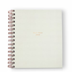 May include: A cream-colored spiral-bound journal with the words "DAILY PAUSE journal" printed in gold. The spiral binding is a copper color. The journal is square and sits on a white background.