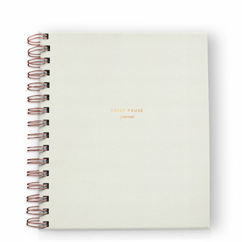 May include: A cream-colored spiral-bound journal with the words "DAILY PAUSE journal" printed in gold. The spiral binding is a copper color. The journal is square and sits on a white background.