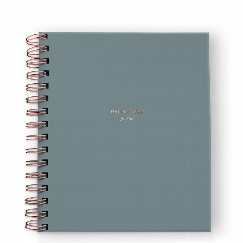 May include: A square, teal-colored journal with a copper-colored spiral binding. The cover features the words "DAILY PAUSE journal" in a gold font. The journal is designed for writing and reflection.