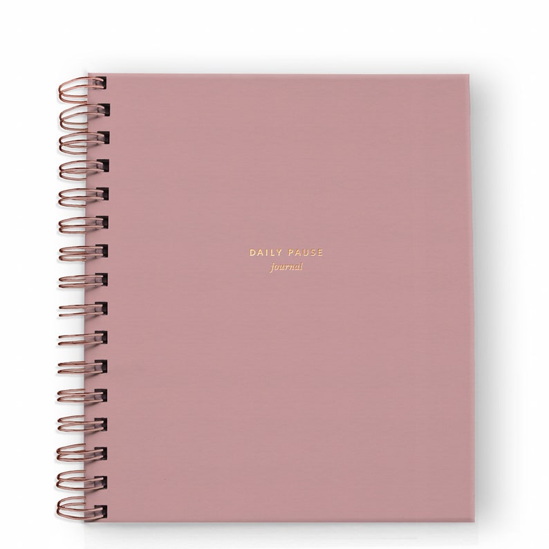 May include: A square, dusty rose journal with a copper spiral binding. The cover features the gold-colored text "DAILY PAUSE journal". The journal is closed and sits on a white background.
