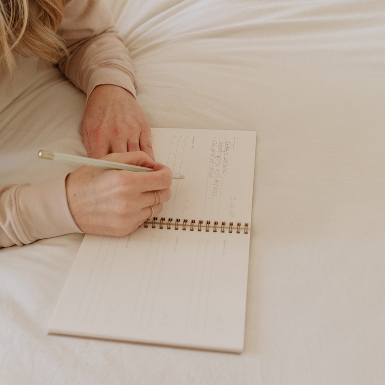 May include: A person writing in a spiral bound notebook with a light green pencil. The notebook is open to a blank page with lines.