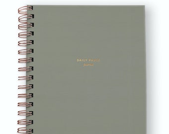 May include: A sage green spiral-bound journal with copper-colored wire binding. The front cover features the text "DAILY PAUSE" in gold capital letters, with the word "journal" printed in a smaller gold script font below it.