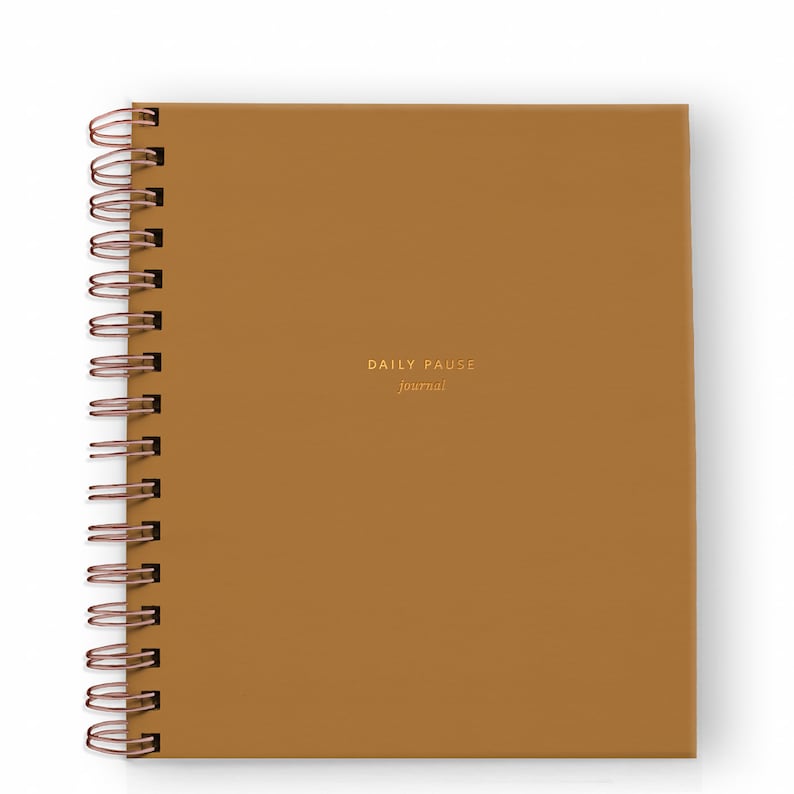 May include: A square, spiral-bound journal with a mustard-colored cover. The words "DAILY PAUSE journal" are embossed in gold on the front. The spiral binding is a copper color.