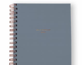 May include: A slate blue spiral-bound planner with copper-colored wire binding. The front cover features the text "DAILY OVERVIEW planner" printed in small, centered copper lettering. The planner has a solid, matte-finish cover.
