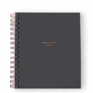 May include: A dark gray, spiral-bound journal with the words "DAILY PAUSE journal" in gold lettering. The spiral binding is a copper color. The journal is square and appears to be made of a textured material.