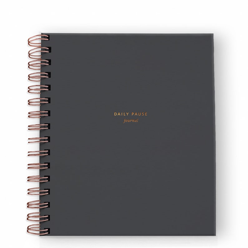 May include: A dark gray, spiral-bound journal with the words "DAILY PAUSE journal" in gold lettering. The spiral binding is a copper color. The journal is square and appears to be made of a textured material.