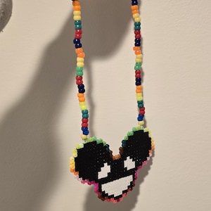 May include: A colorful beaded necklace with a pixelated black and white image of a cartoon face. The face has a large smile and two small eyes.