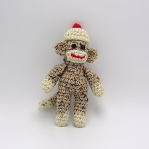 May include: A crocheted sock monkey wearing a white crocheted hat with a red pom-pom. The monkey has brown and white yarn and a red embroidered smile.