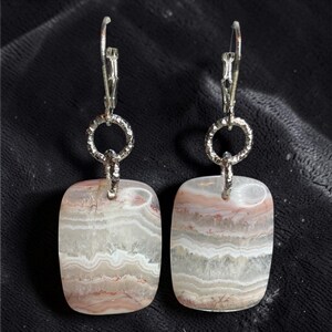 May include: A pair of drop earrings featuring rectangular, banded agate stones with wavy layers of white, gray, and soft pink. Each stone hangs from a textured, circular silver-toned metal link attached to a silver-toned metal lever-back earring hook.