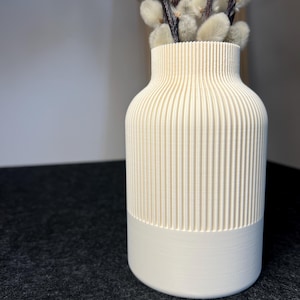 Könnte beinhalten: Eine cremefarbene Vase mit vertikalen Rippendetails und einem glatten, soliden Sockel. Die Vase enthält ein Bündel flauschiger, hellfarbiger Kätzchenzweige. Die Vase ist ein dekorativer Artikel für die Inneneinrichtung.