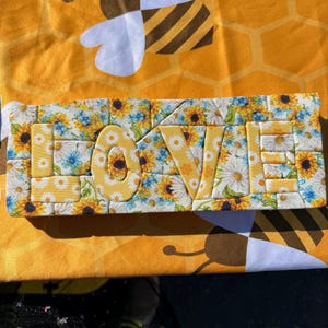 May include: A rectangular decorative sign featuring the word LOVE in raised, padded letters. The surface is covered in a fabric pattern of yellow sunflowers, white daisies, and small blue flowers against a light background, creating a floral mosaic effect.