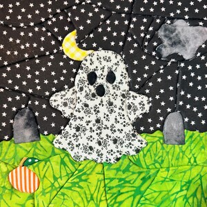 May include: A fabric quilt block featuring a white ghost with a black floral pattern, centered against a black sky with white stars. The scene includes a yellow crescent moon, two gray tombstones, a green grassy field, and an orange striped pumpkin.
