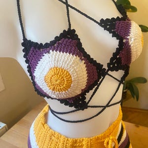 Può includere: Un set di bikini in crochet composto da top e slip, nei colori viola, bianco, giallo e nero. Il top presenta un cerchio in crochet con un centro giallo e lo slip ha un motivo a righe.