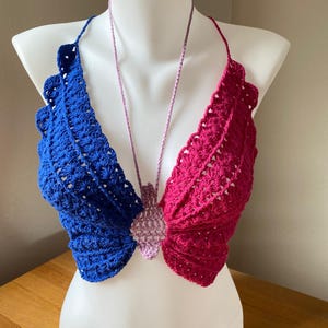 Puede incluir: Un top de ganchillo hecho a mano con diseño de alas de mariposa. El top presenta alas azules y magenta con un centro lavanda. El top halter se muestra en un maniquí blanco. El ganchillo tiene un aspecto texturizado.