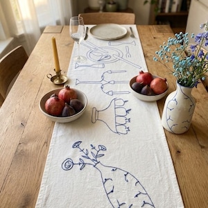 May include: A white linen table runner featuring blue embroidered line art designs of tableware, a candelabra, and vases with flowers. The runner is laid on a wooden table with bowls of pomegranates, a candle, a wine glass, and a ceramic vase.