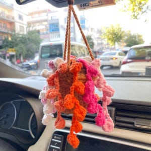 Peut inclure: Un porte-plante en crochet fait à la main, suspendu dans une voiture. Le pot marron est rempli de feuilles en crochet colorées, dans des tons orange, rose et blanc. Le support est suspendu au rétroviseur par une corde marron et une perle en bois.