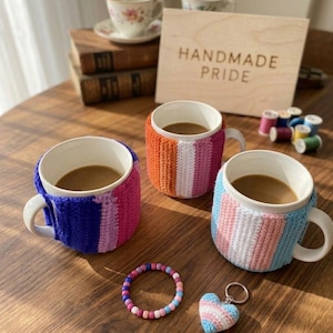 Könnte beinhalten: Drei weiße Keramikbecher mit Kaffee, jeder in einer andersfarbigen Häkelhülle. Ein Holzschild mit der Aufschrift "HANDMADE PRIDE". Ein Perlenarmband und ein herzförmiger Schlüsselanhänger sind ebenfalls zu sehen.