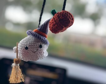 Puede incluir: Encanto de coche de Halloween tejido a ganchillo. Presenta un fantasma blanco con un sombrero de bruja con detalles morados y naranjas, sosteniendo una pequeña escoba. Una calabaza naranja tejida a ganchillo con un tallo verde cuelga cerca.