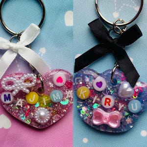 May include: Two heart-shaped keychains with glitter and embellishments. One is pink with white pearls and the letters M, A, J, and M. The other is purple with a black bow and the letters K, I, R, Y, and U. Both have key rings.