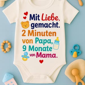 May include: White baby onesie with the text "Mit Liebe, gemacht. 2 Minuten von Papa, 9 Monate von Mama." in various colors. The onesie has a bear and heart graphic. Baby accessories surround the onesie.