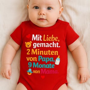May include: A baby wearing a red onesie with the text "Mit Liebe, gemacht. 2 Minuten von Papa, 9 Monate von Mama." The onesie has a teddy bear, heart, bottle, and pacifier graphic. The baby is wearing a pink bow headband.