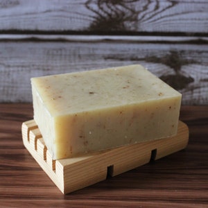 May include: A bar of natural soap with a light beige color and a grainy texture, resting on a wooden soap dish.