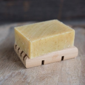 May include: A bar of natural soap on a wooden soap dish. The soap is a light yellow color and has a textured surface. The soap dish is made of light brown wood and has a rectangular shape.
