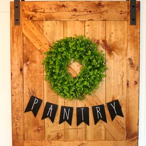 May include: A wooden door with a black metal handle and a black and white banner that says "PANTRY" hanging from it. A green wreath is hanging on the door.