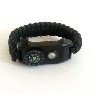 May include: Black paracord bracelet with a built-in compass and a small flashlight. The compass has a green needle and white markings.