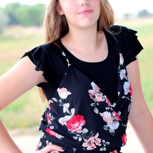 May include: A woman wearing a black sleeveless top with a floral print overlay. The top has a V-neckline and a ruffled trim on the shoulders.