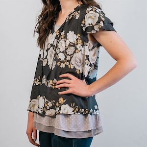 May include: A black floral print blouse with a ruffled neckline and short sleeves. The blouse has a layered look with a gray undershirt.