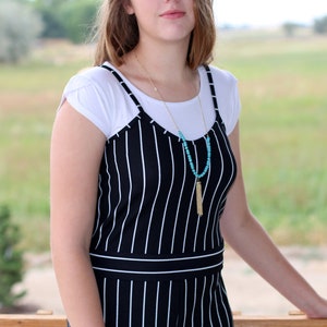 May include: A woman wearing a white t-shirt layered under a black and white striped jumpsuit. She is wearing a gold chain necklace with a turquoise beaded tassel.