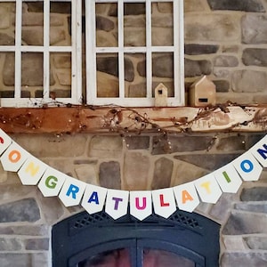 Puede incluir: Una pancarta blanca con la palabra "CONGRATULATIONS" escrita en colores arcoíris cuelga sobre una chimenea.