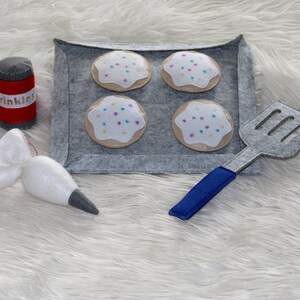 May include: Felt play food set with four cookies on a baking sheet, a piping bag, and a spatula. The cookies are decorated with white frosting and colorful sprinkles. The container says "Sprinkles".